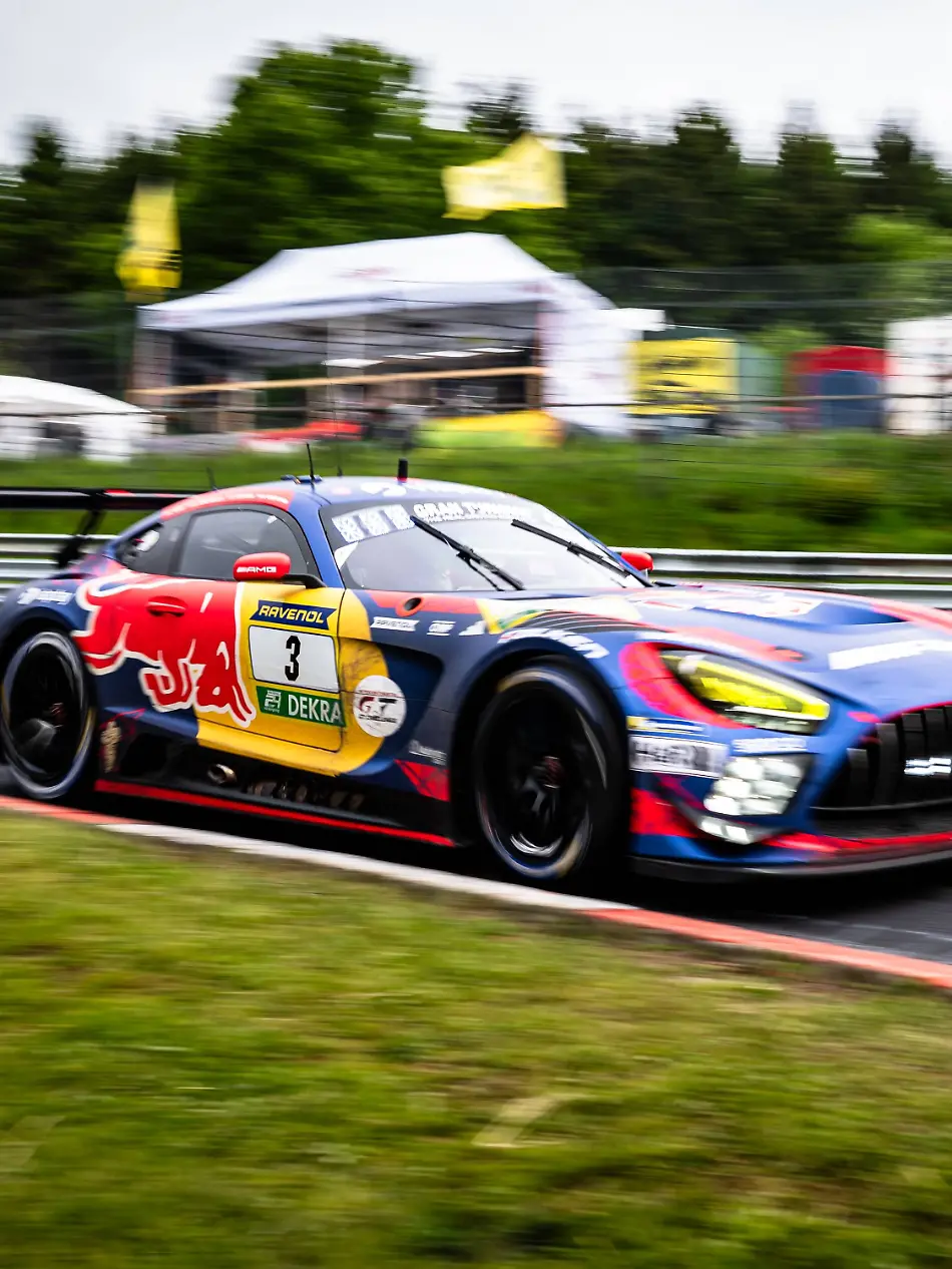 Der-Mercedes-AMG-GT3-mit-der-Startnummer-3-von-Mercedes-AMG-Team-Verstappen-Racing-durchfaehrt-mit-hoher-Geschwindigkeit-eine-Kurve-auf-dem-Nuerburgring-Das-Fahrzeug-traegt-das-markante-Red-Bull-Livery-in-Blau-Rot-und-Gelb-mit-RAVENOL-und-DEKRA-Sponsoring-Die-Panning-Aufnahme-betont-die-Dynamik-des-Rennwagens-waehrend-der-Hintergrund-mit-Boxenanlagen-und-Baeumen-verschwimmt