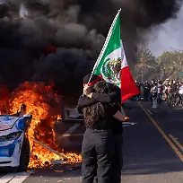Anti-ICE-Protests-in-Los-Angeles-A-protestor-waves-a-Mexican-flag-in-front-of-a-burning-car-during-a-demonstration-in-Los-Angeles-California-on-June-8-2025-Protests-broke-out-across-Los-Angeles-in-response-to-federal-immigration-raids-and-the-deployment-of-National-Guard-troops-by-the-Trump-administration-The-protests-led-by-immigrant-rights-groups-and-local-residents-quickly-escalated-into-street-clashes-with-law-enforcement-Demonstrators-waved-Mexican-flags-held-signs-against-ICE-and-set-fire-to-cars-in-acts-of-defiance